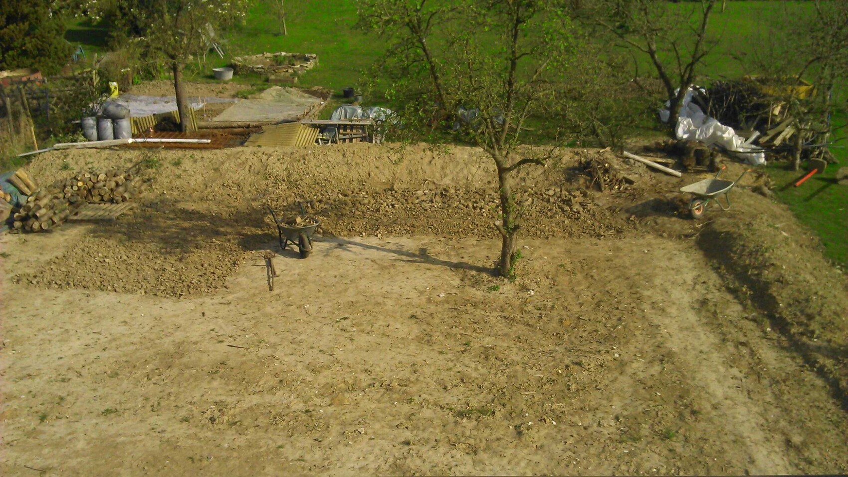 Baustelle im Garten mit Erdehaufen, Schubkarre, Holzstapeln und Baum auf unebenem Boden