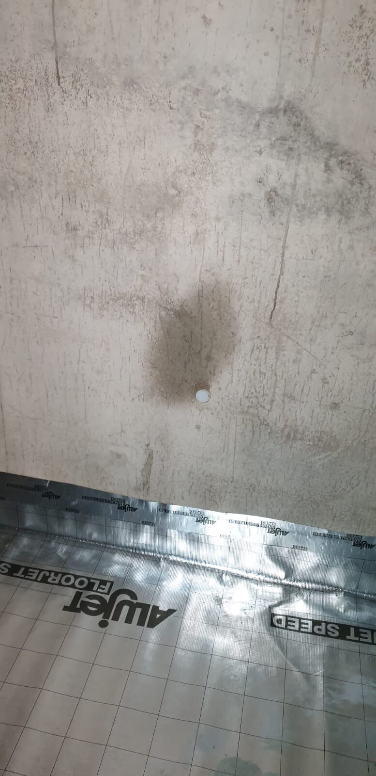 Betonwand mit Flecken; kleine weiße runde Abdeckung; darunter Folie auf Fliesenboden.