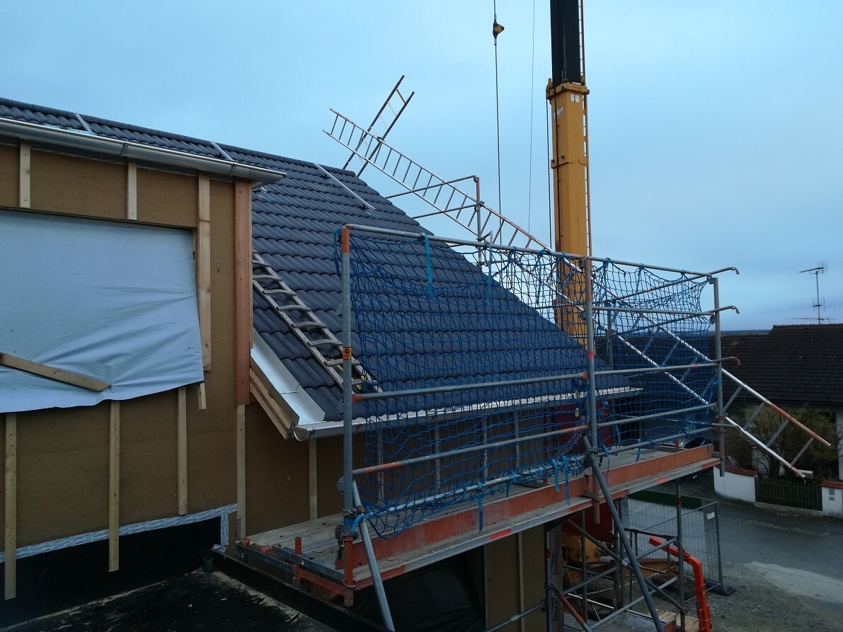 Dachbaustelle eines Hauses: Gerüst, blaue Sicherheitsnetze, Kranarm und Dacharbeiten.