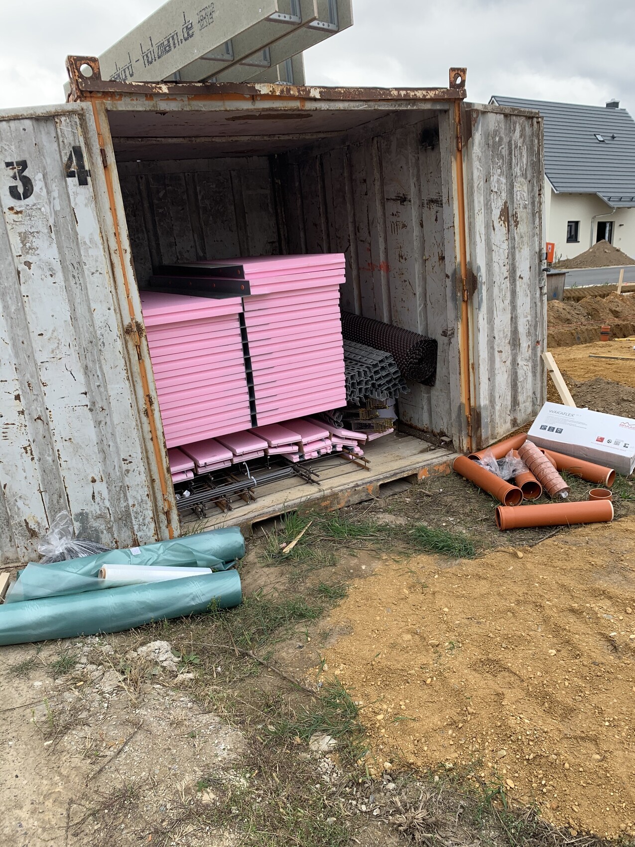 Offener Versandcontainer mit pinken Dämmplatten, Rolldraht und Rohren auf Baustelle.