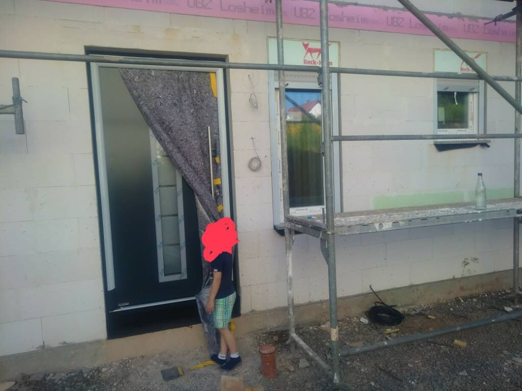 Ein Kind steht vor einer offenen Tür auf einer Baustelle mit Gerüst und Fenster.