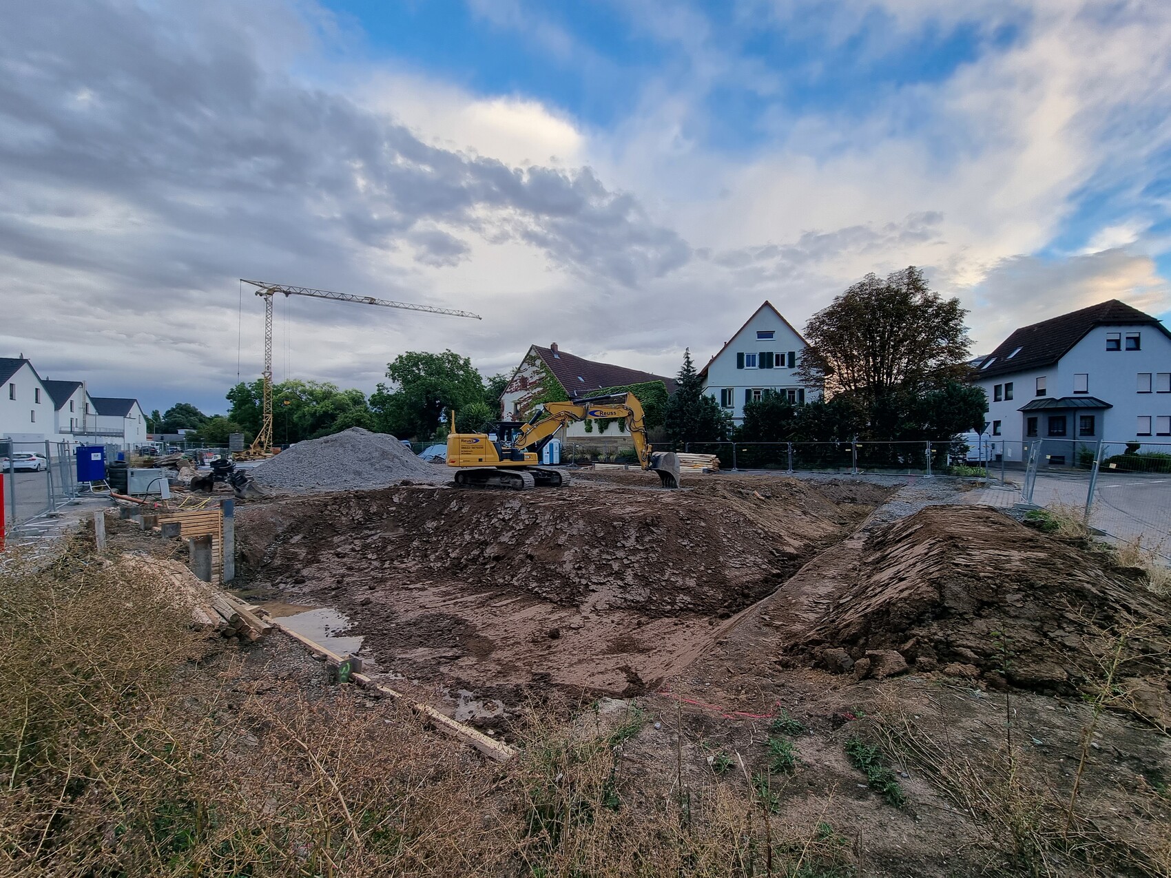 Große Baugrube in Wohngegend; Bagger, Kran, Erdmassen, Häuser im Hintergrund