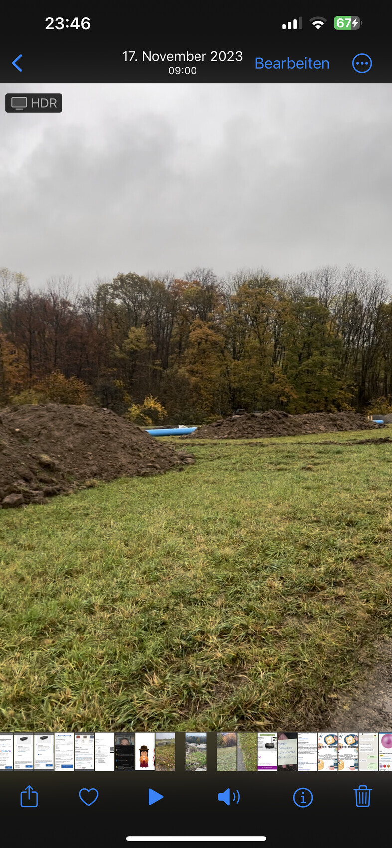 Grünes Feld mit Erdhaufen, blauen Rohren, Herbstbäumen im Hintergrund unter grauem Himmel.