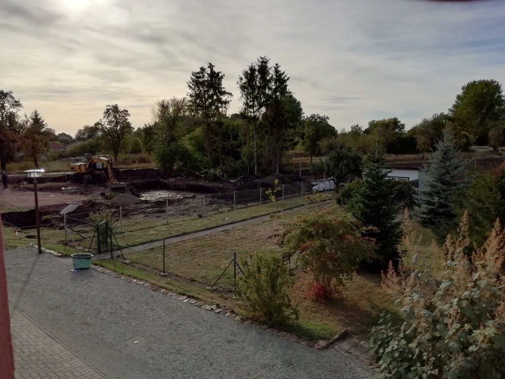 Baustelle mit Bagger, Grabenarbeiten, Zaun und umliegendem Garten.