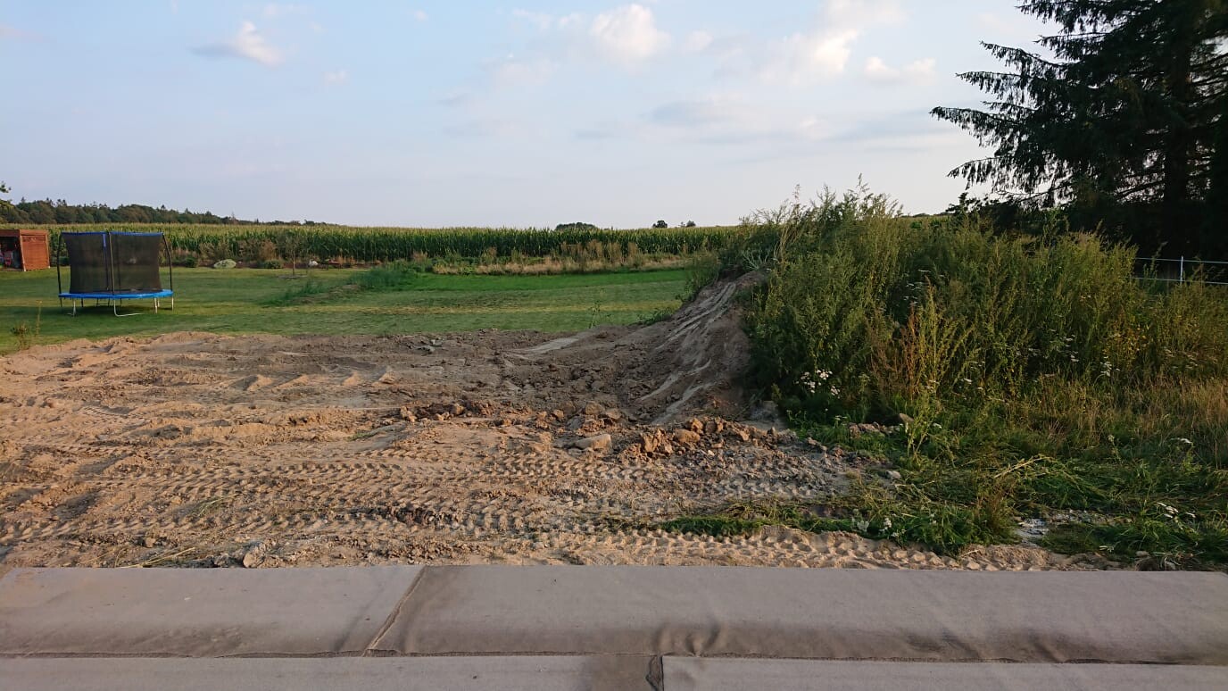 Baustelle im Freien mit Erdarbeiten, unbefestigtem Boden, Grasfläche und Trampolin im Hintergrund