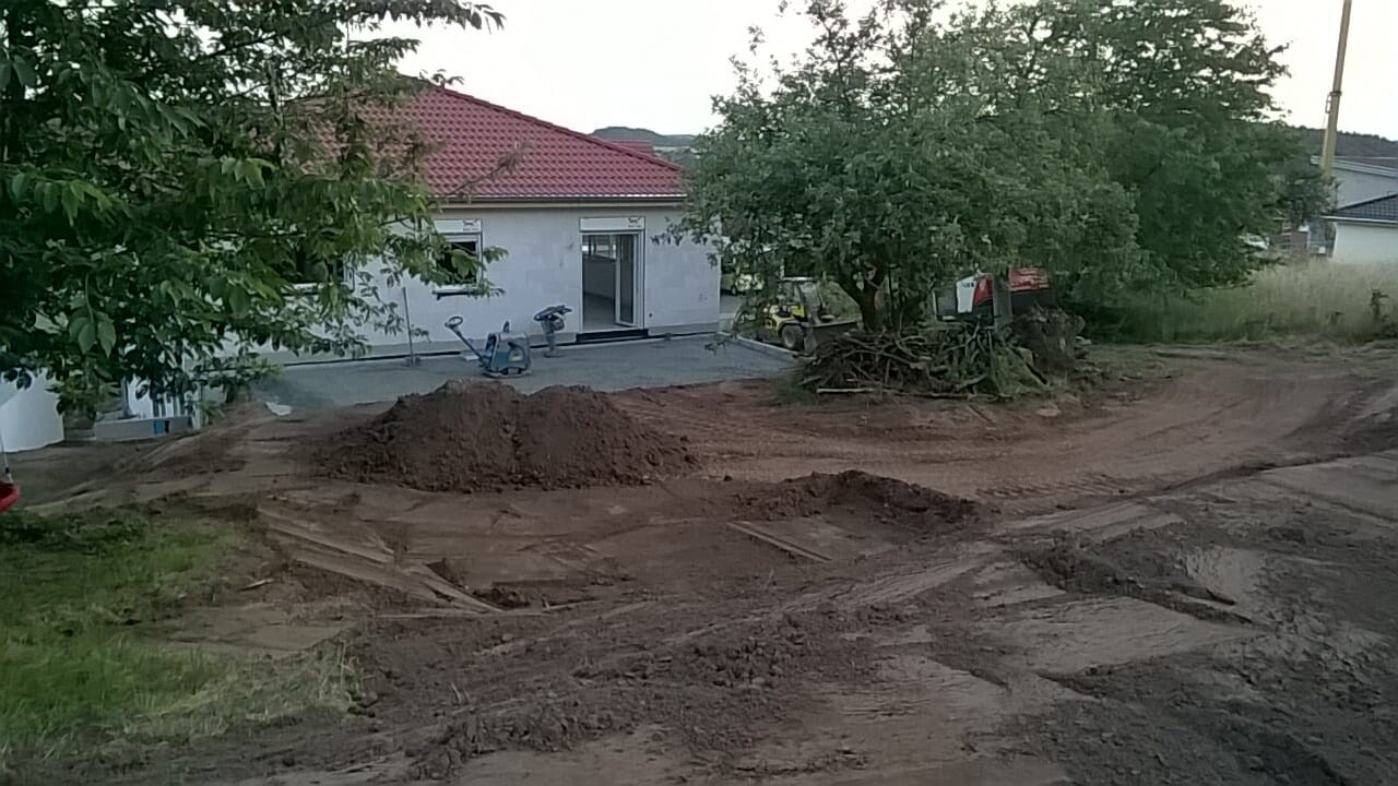 Baustelle vor Haus: Erdaushub, Dreckhügel, Baugeräte und Bäume sichtbar.