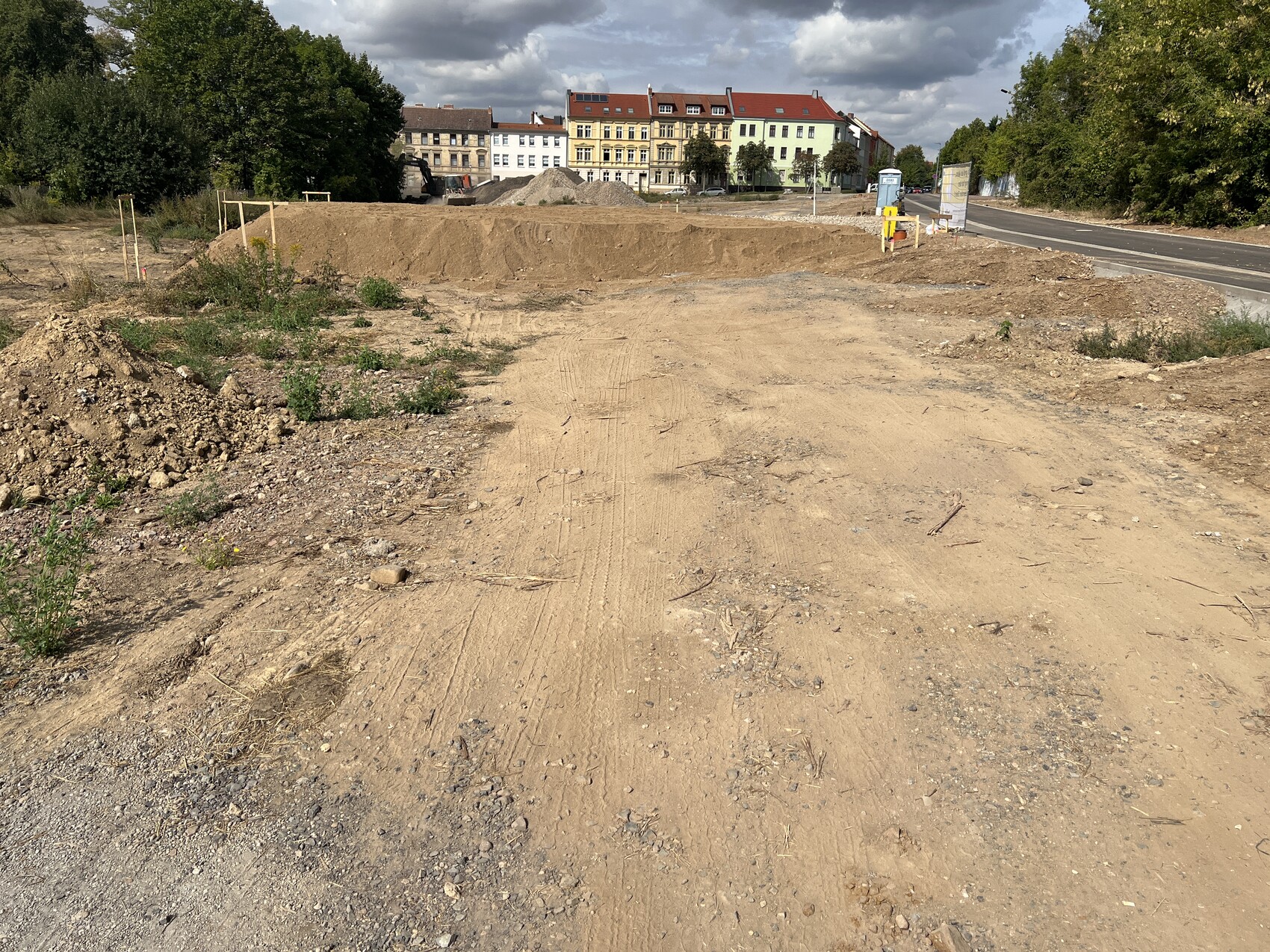 Vordergrund unbefestigte Baustelle mit Erdhügeln, Kies und Boden; Häuserreihe im Hintergrund.