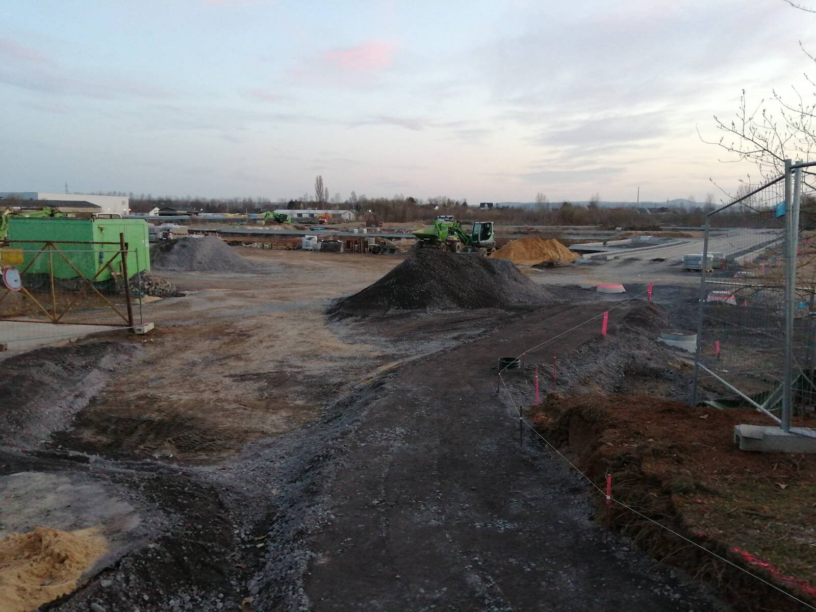 Baustelle mit Kies- und Erdhügeln, grünen Containern, Baumaschinen im Hintergrund