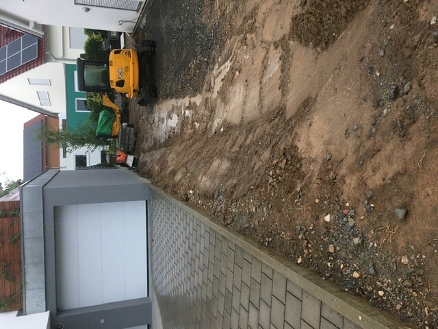 Kleiner gelber Minibagger auf Baugrund vor Haus mit Garage; Pflasterweg und Erde sichtbar.