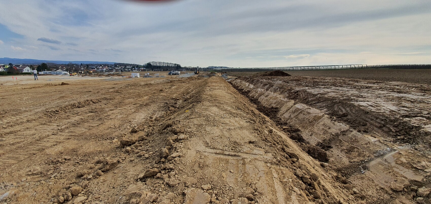 Große Baustelle mit Gräben, Erdmassen und Baufahrzeugen; Dorf und Felder im Hintergrund.