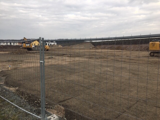Baustelle: Bauzaun im Vordergrund, unbefestigte Erde, zwei gelbe CAT-Bagger im Hintergrund.