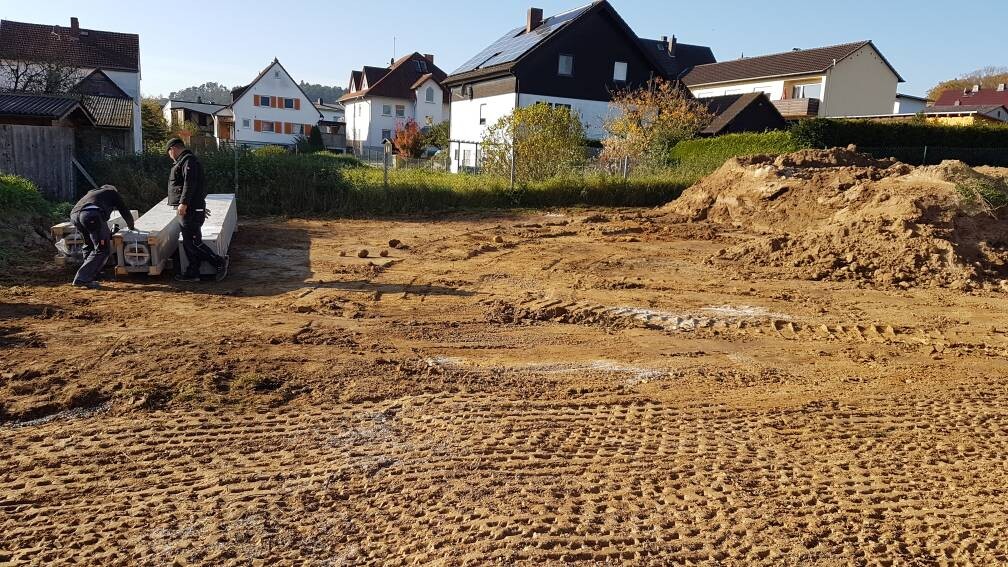 Baustelle in Wohngegend: zwei Arbeiter legen Betonblöcke, Erde und Baugeräte sichtbar.