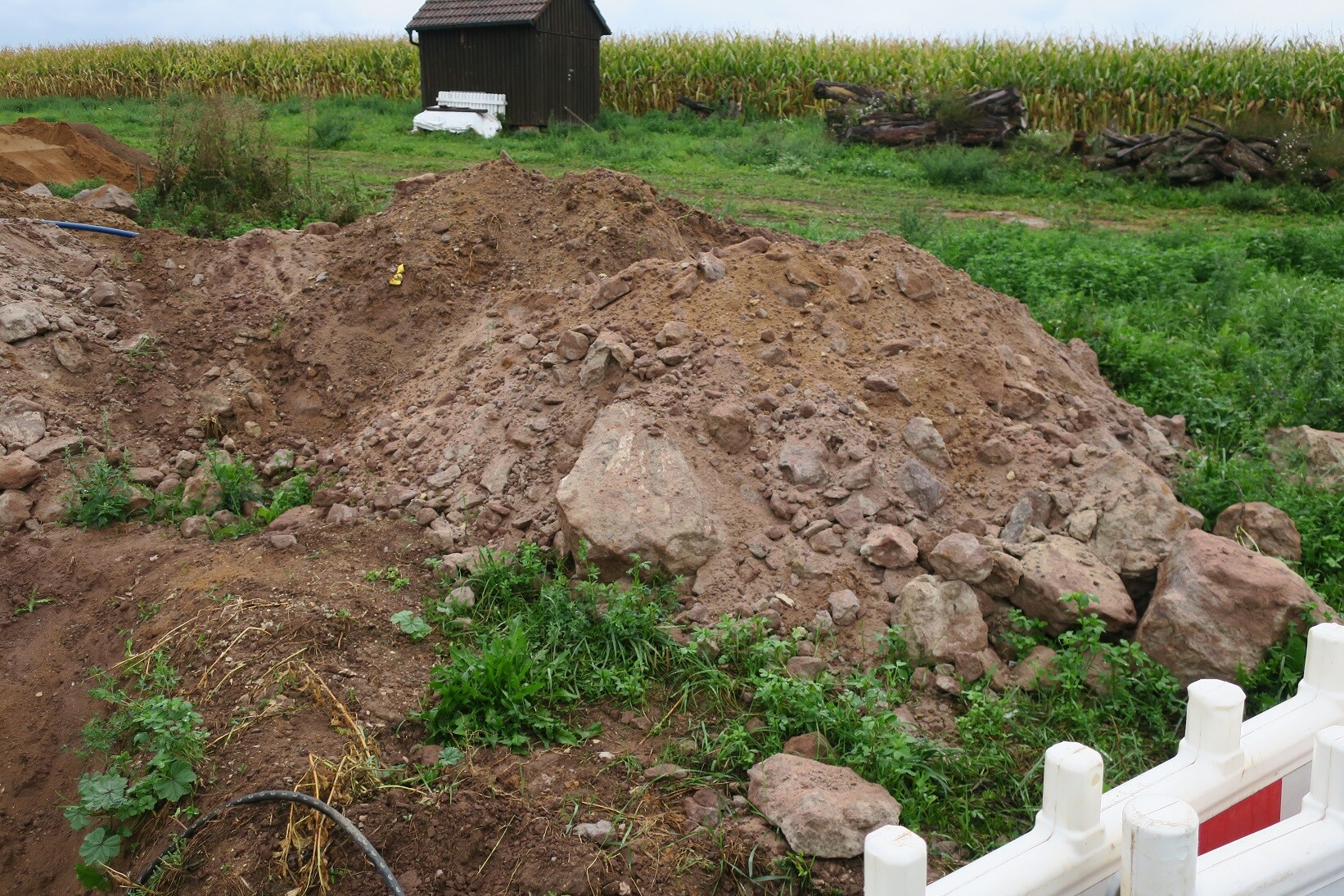 Große Erdmassen, Felsbrocken und Baugerät auf einer Baubaustelle mit Feld im Hintergrund.