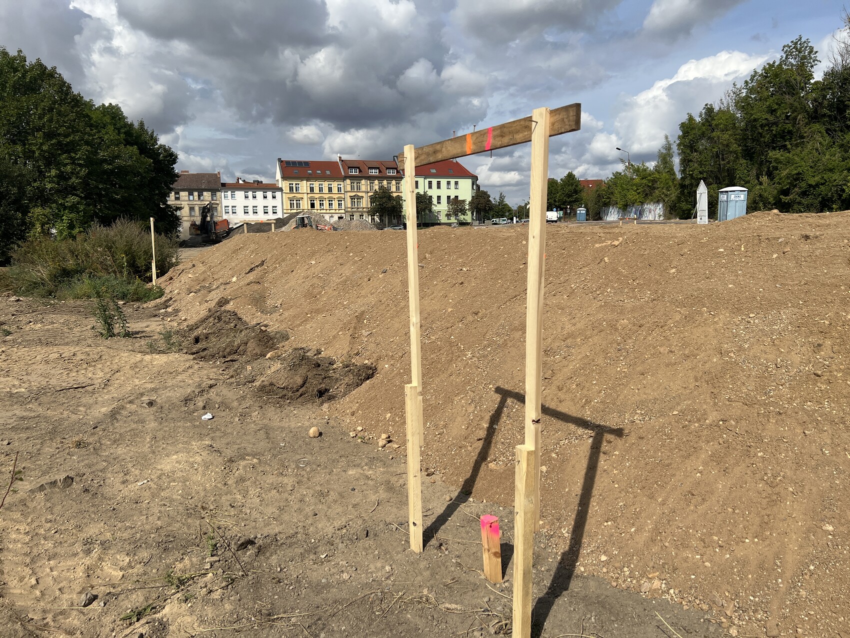 Baufläche mit Holzgestell im Vordergrund, Erdhügeln und Häusern im Hintergrund.
