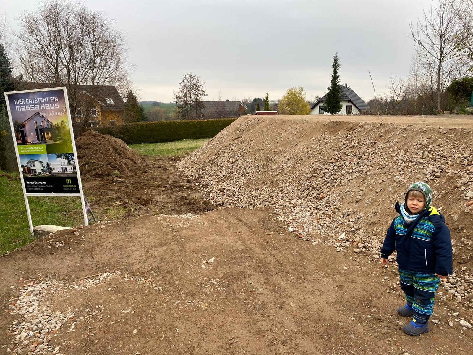 Ein Kind in Winterkleidung steht vor einem Erdhügel/Kieshügel auf einer Baustelle; Schild links.