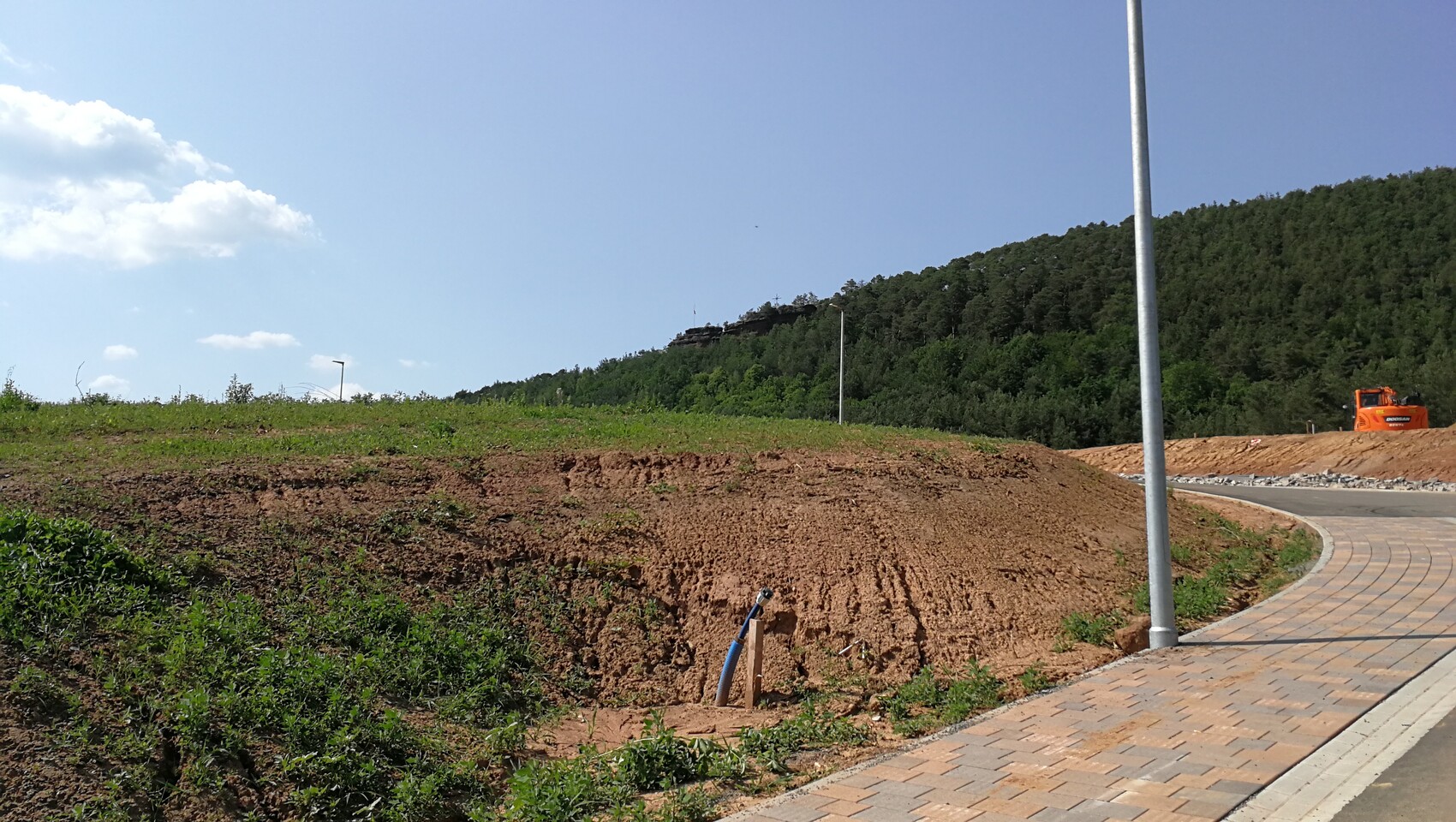 Gepflasterter Kurvenweg, Erdhügel, blaue Rohrleitung, Laternenmast, orange Baumaschine, Wald.