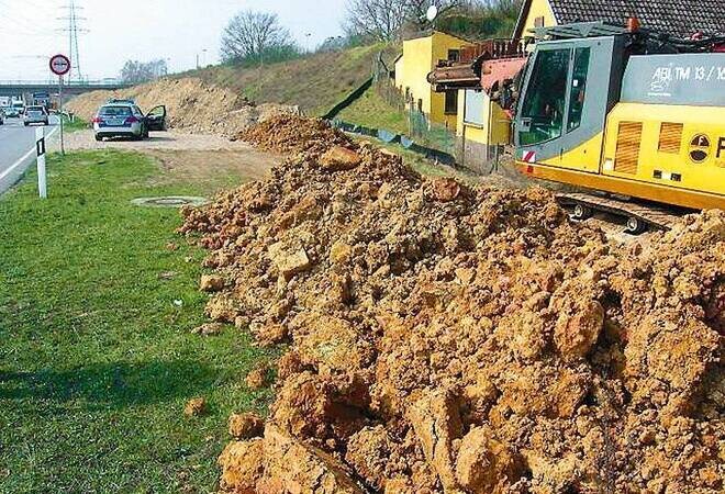 Großer Erdhügel am Straßenrand neben Bagger und Haus dahinter.