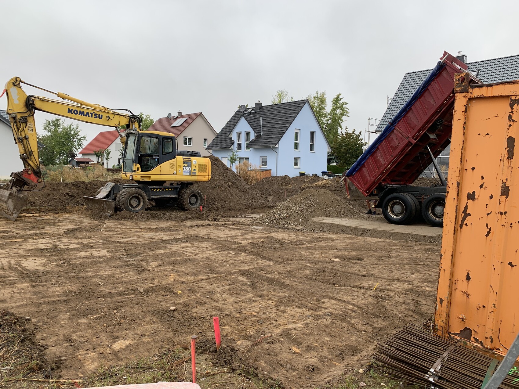 Gelber Komatsu-Bagger lädt Erde auf einen roten Kipp-LKW auf einer Baustelle, Häuser im Hintergrund.