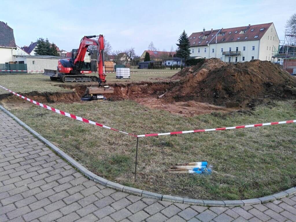 Roter Minibagger gräbt Graben auf Grünfläche vor Wohnhäusern; Absperrband, Erdaushub.