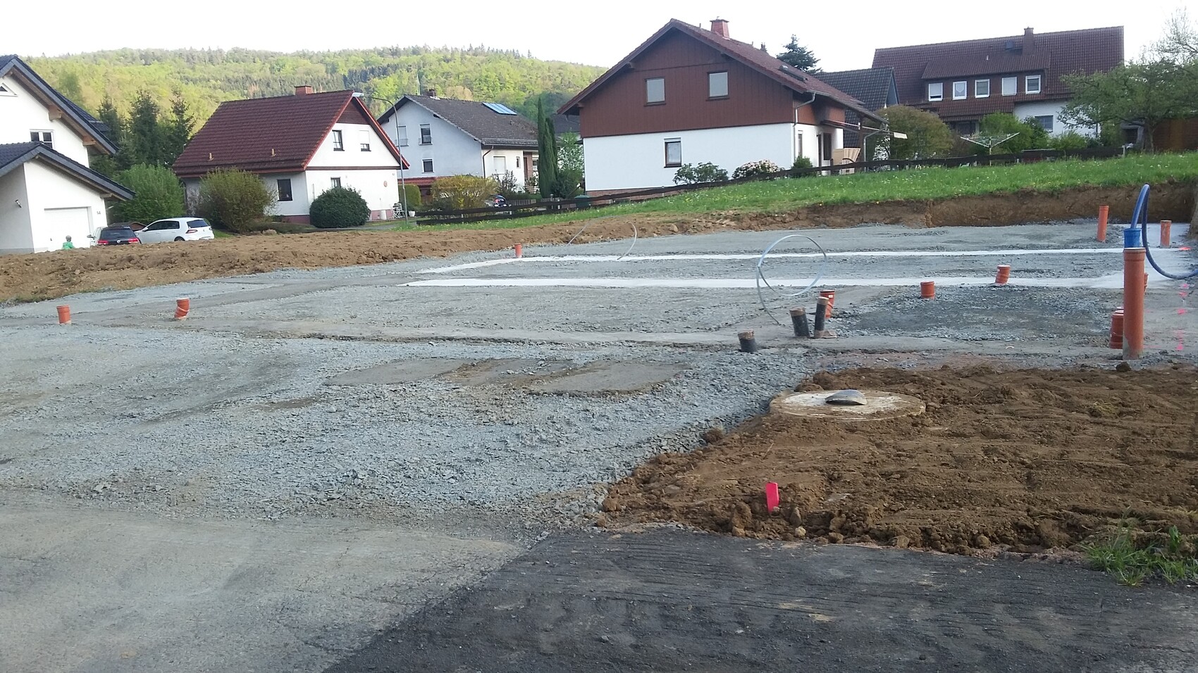 Wohngebiet: Kiesuntergrund, Rohre ragen aus dem Boden, Häuser im Hintergrund.