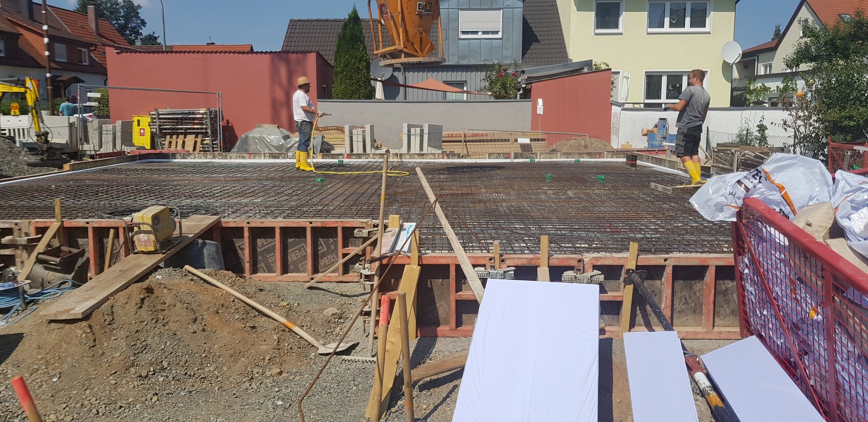 Baustelle: Arbeiter mit gelben Stiefeln stehen auf Bewehrung eines Fundaments; Häuser im Hintergrund.