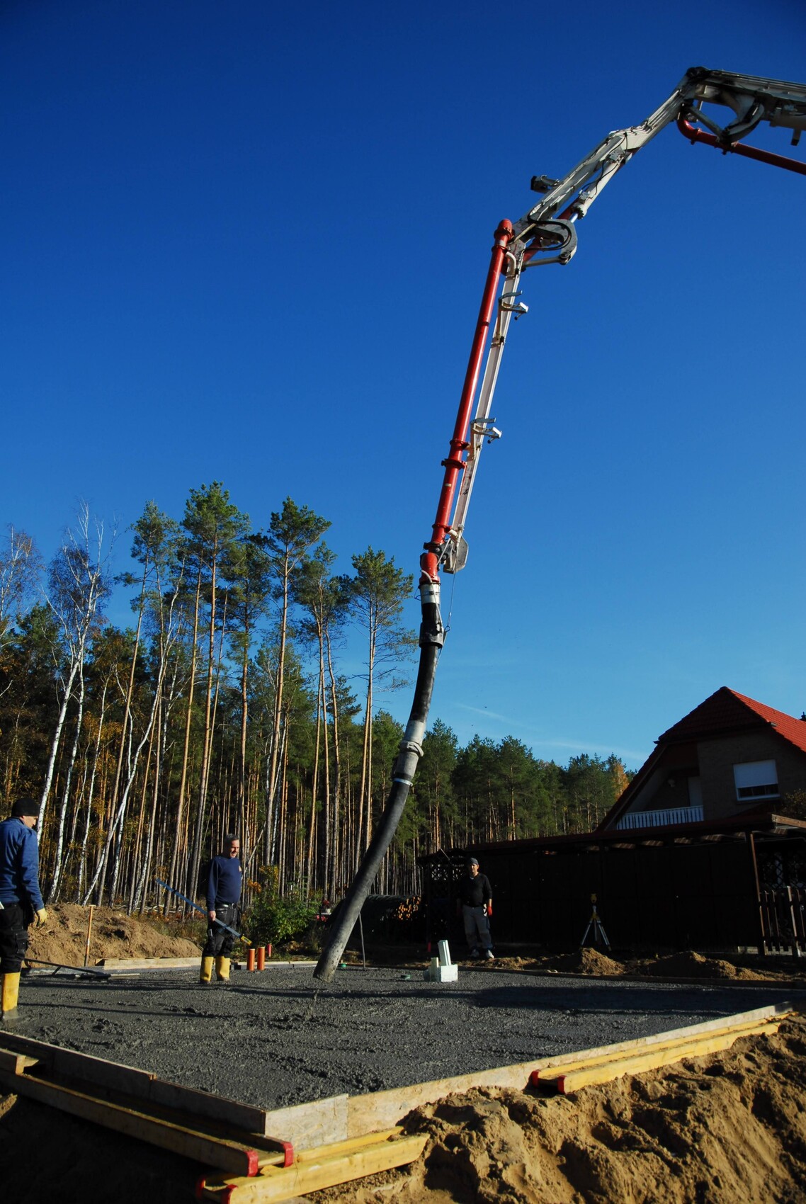 Bauarbeiter gießen Beton durch Schlauch auf Fundament; Kran mit Pumpe, Wald und Haus im Hintergrund