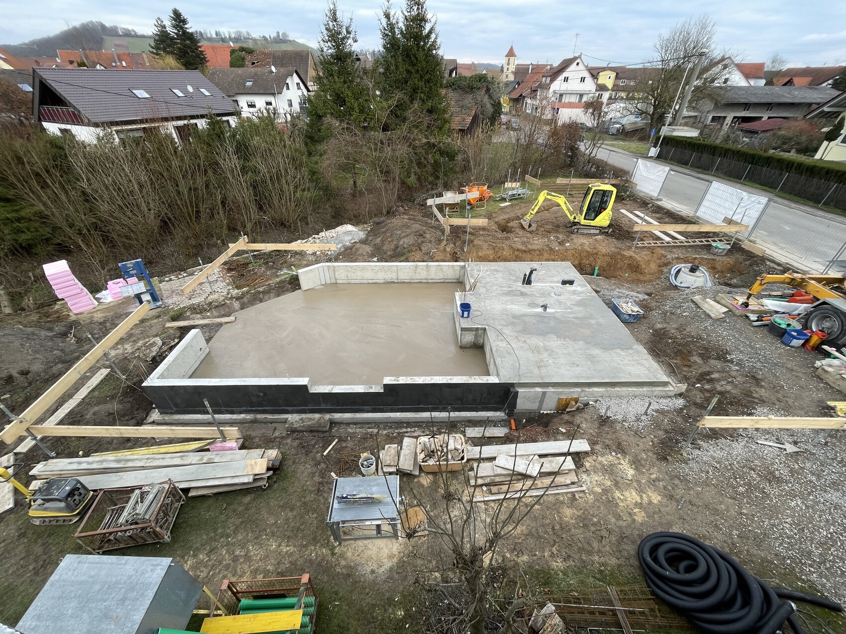 Baustelle mit Fundamentplatte, Bagger, Holzbohlen; Wohngebiet im Hintergrund.