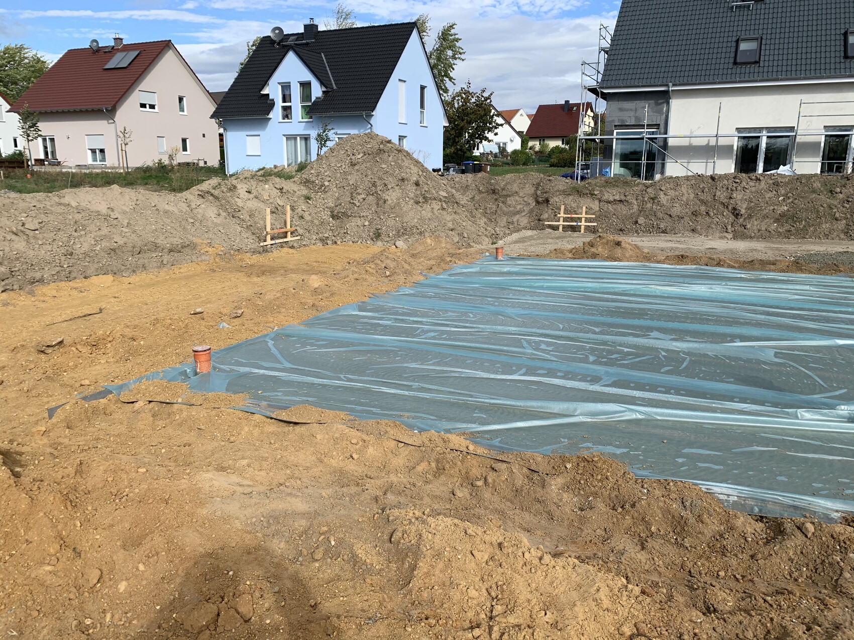 Baustelle: Grube mit blauer Folie, umliegende Erdmassen, Häuser im Hintergrund.