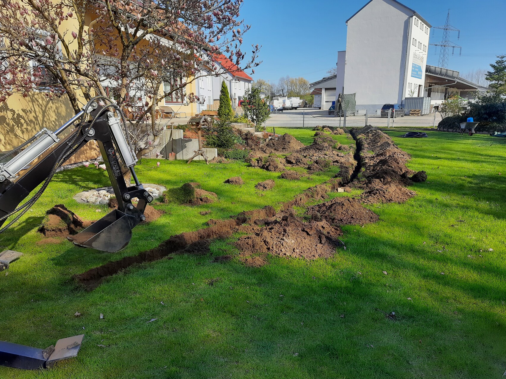 Bagger gräbt eine lange Grube im Gras des Gartens; Erdhaufen säumen die Grabung.