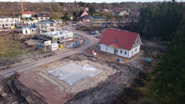 Baustelle mit Fundamentplatte im Vordergrund, rotes Dach und Neubaugebäude im Hintergrund; Kran.