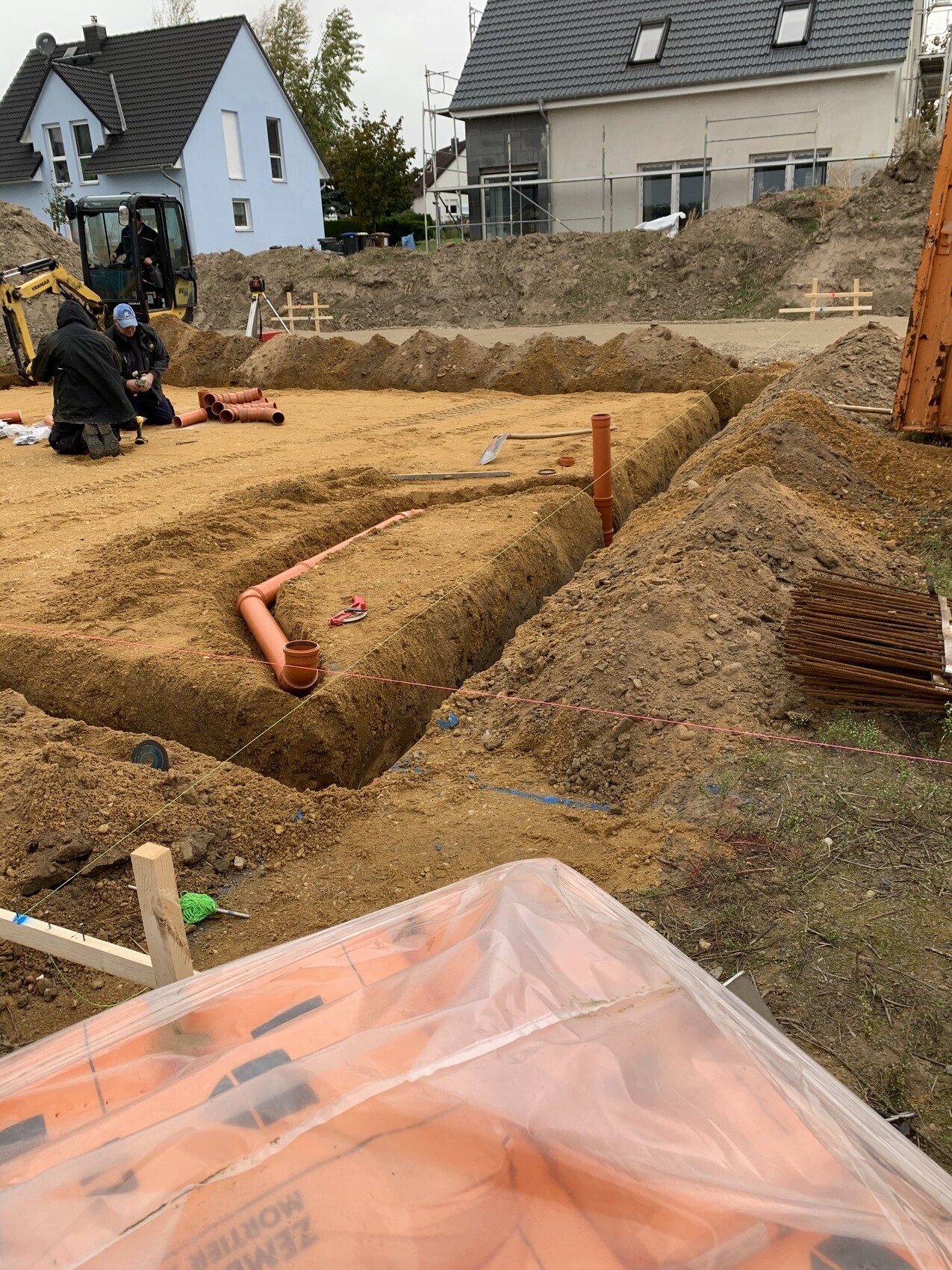 Baustelle: Gräben, orange PVC-Rohre werden verlegt; Arbeiter arbeiten am Boden.
