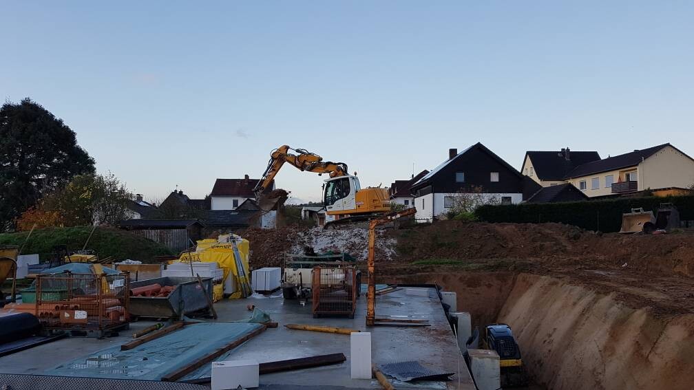 Baustelle mit Bagger auf Erdhügel, Baumaterialien und Wohnhäuser im Hintergrund.