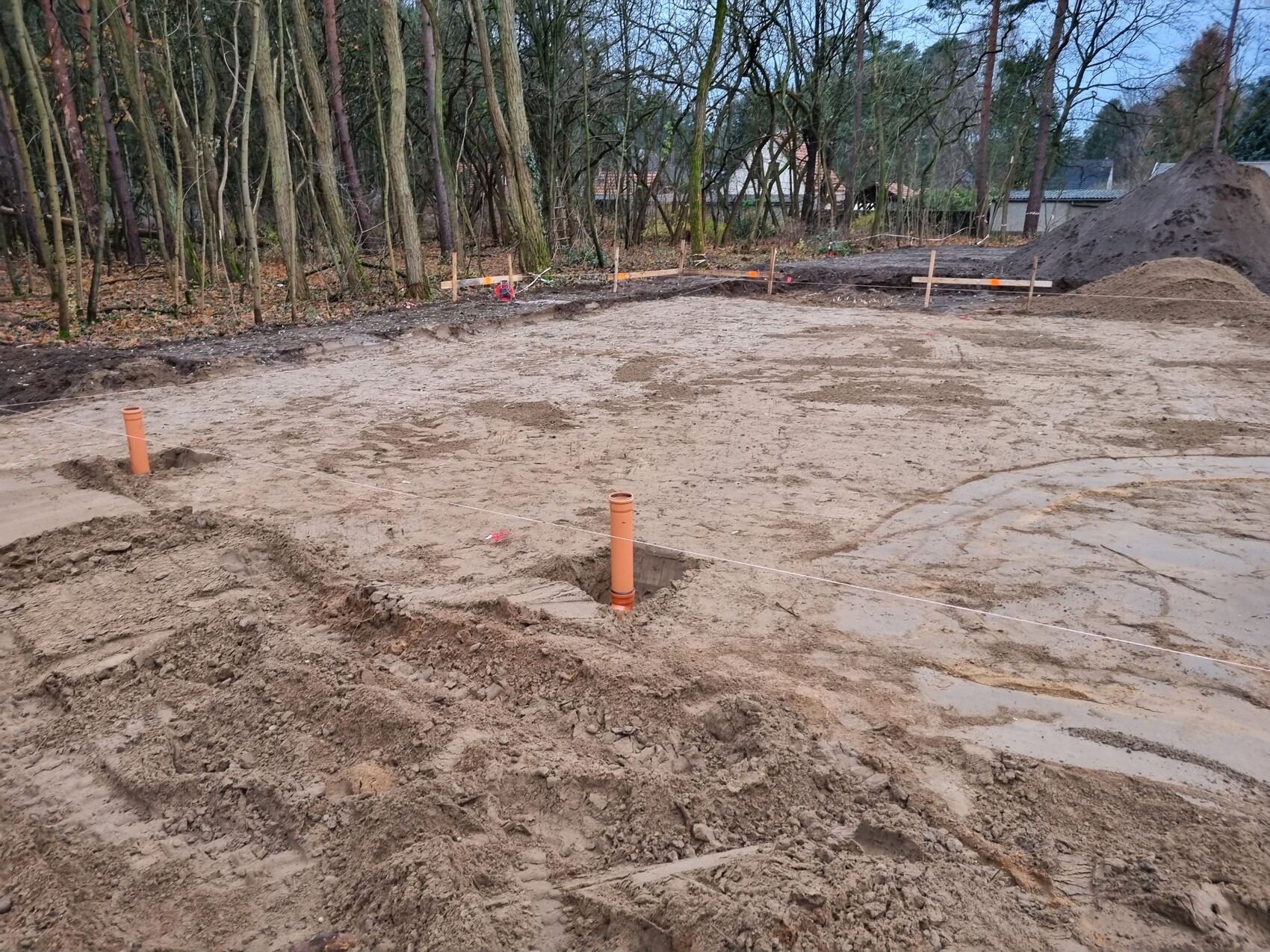 Baustelle mit Erde, orange PVC-Rohre, Baugrube, Erdanhäufungen und Wald im Hintergrund.