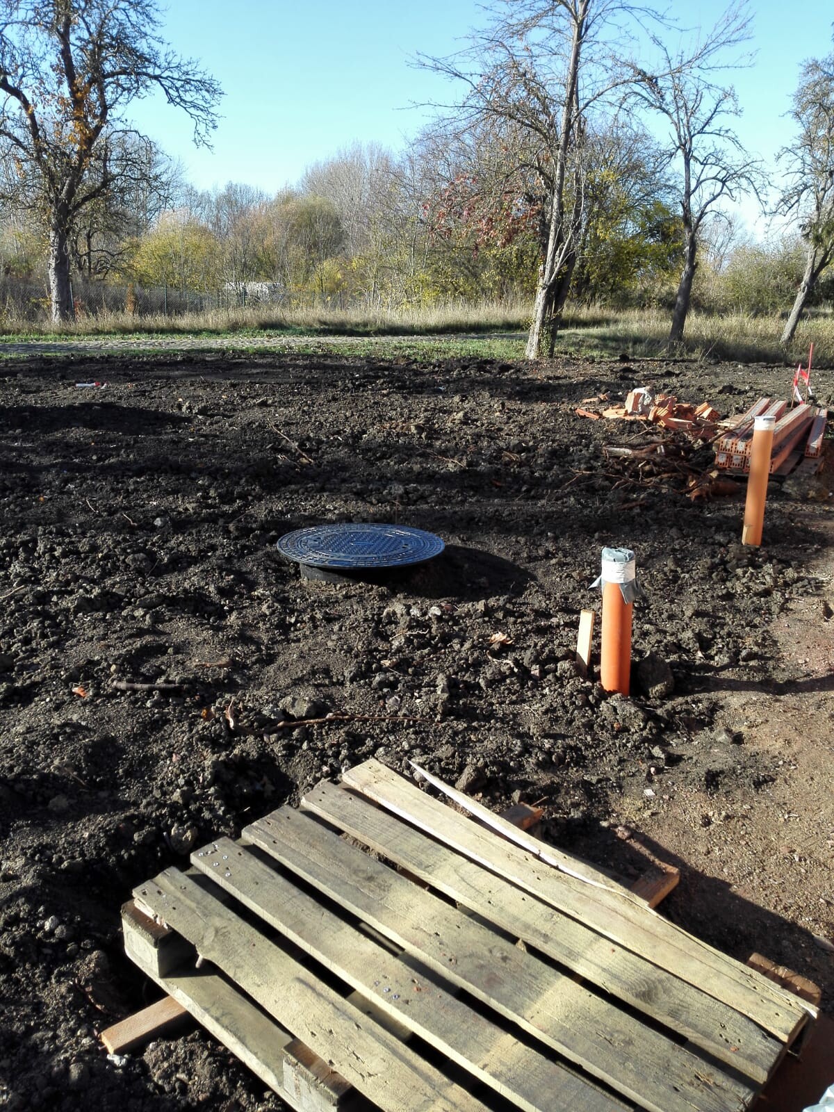 Baufläche im Freien: Erdarbeiten, Holzpaletten, orange Rohre, Bruchstücke, Bäume im Hintergrund.