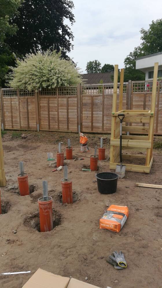 Gartenbaustelle mit orange Rohre in Löchern, Holzgerüst, Eimer, Zementsack.