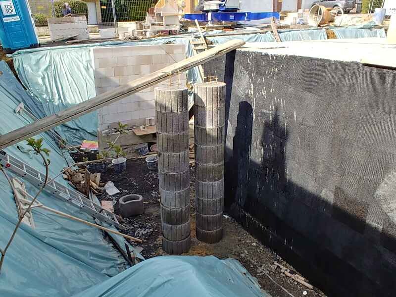 Zwei vertikale Betonstützen stehen in einer Baugrube, umgeben von Planen und Bauequipment.