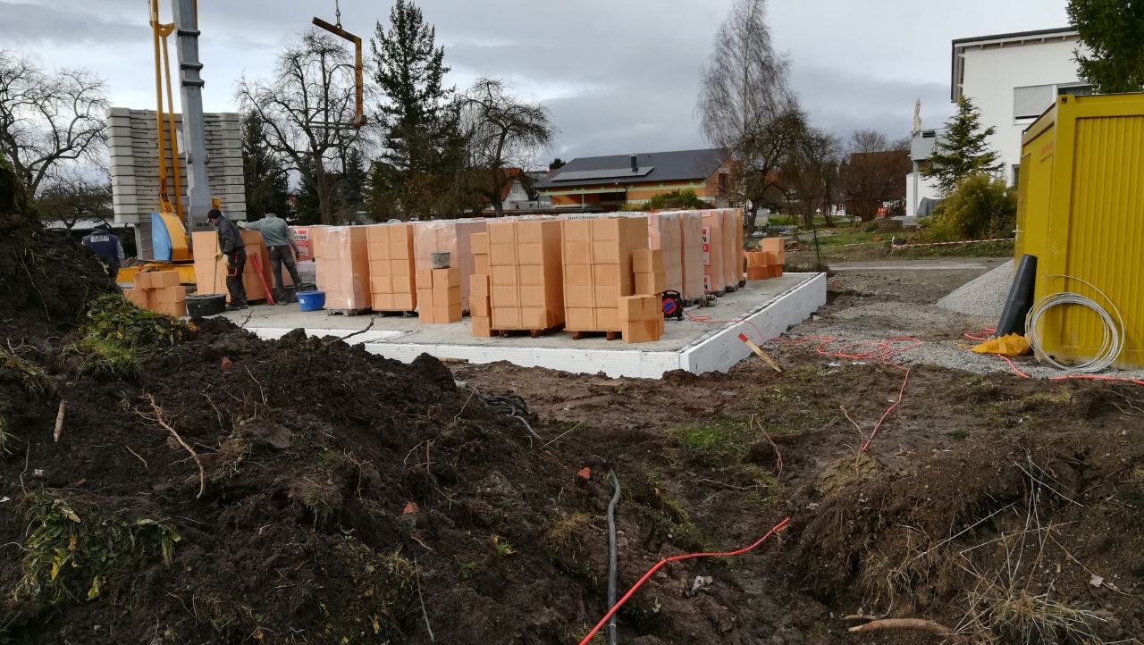 Baustelle mit Fundamentplatte, gestapelten Ziegelblöcken, Bauarbeitern, Gelbcontainer