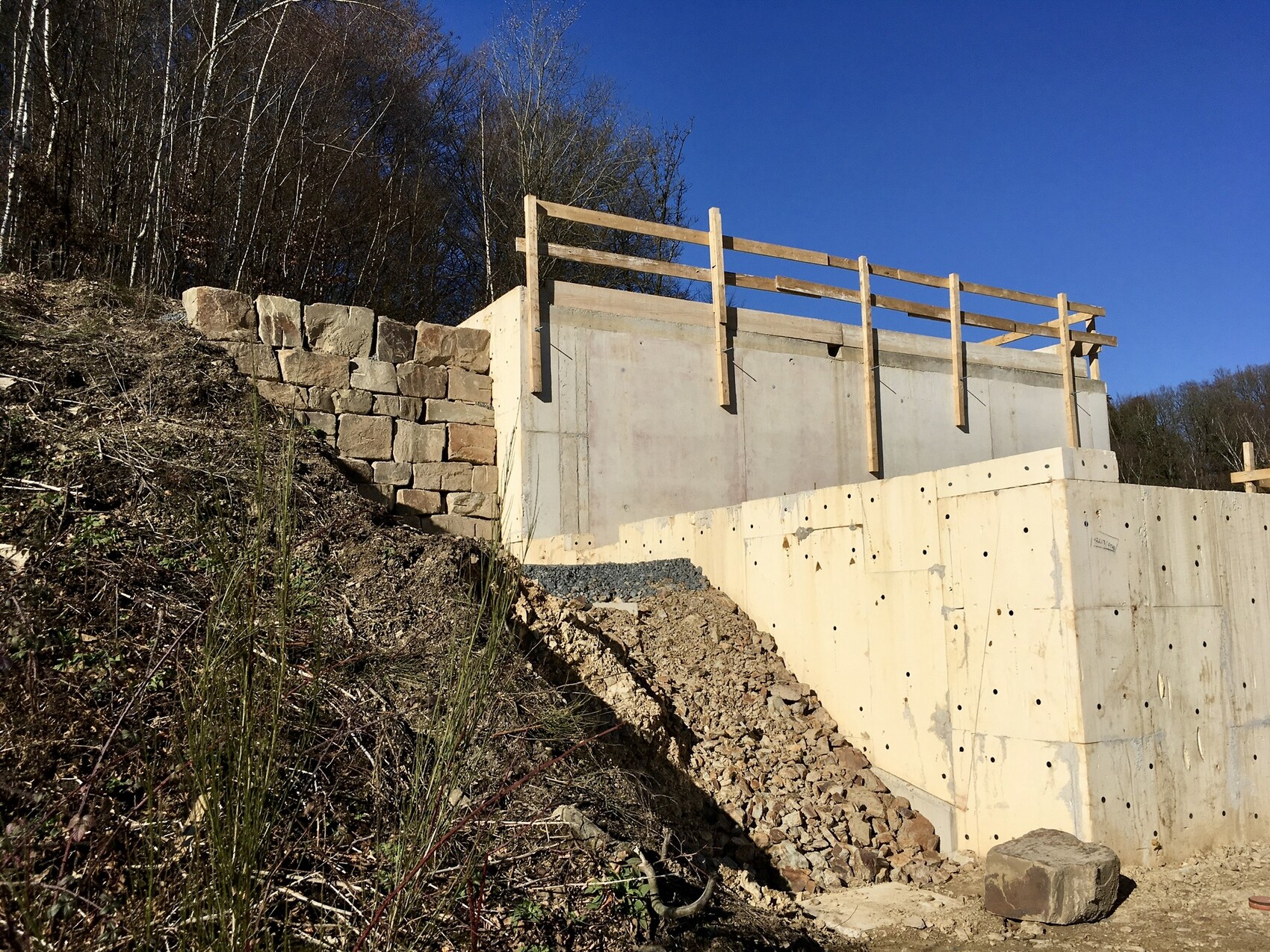 Betonwand mit Holzgeländer am Hang, linke Natursteinmauer und Baugeröll; blauer Himmel.