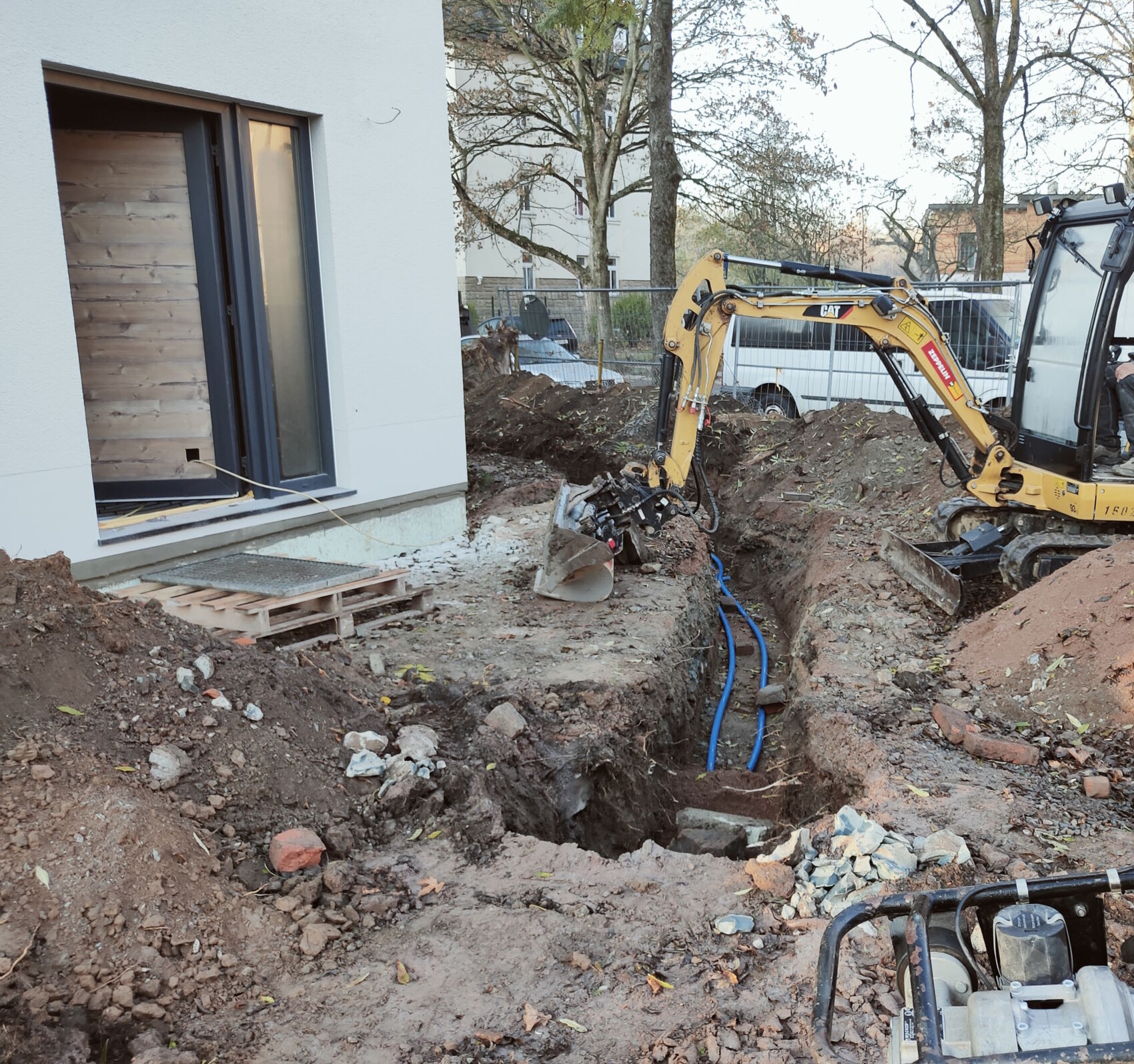 Gelber Minibagger gräbt eine Baugrube neben einem Haus; blaue Rohre liegen im Boden.