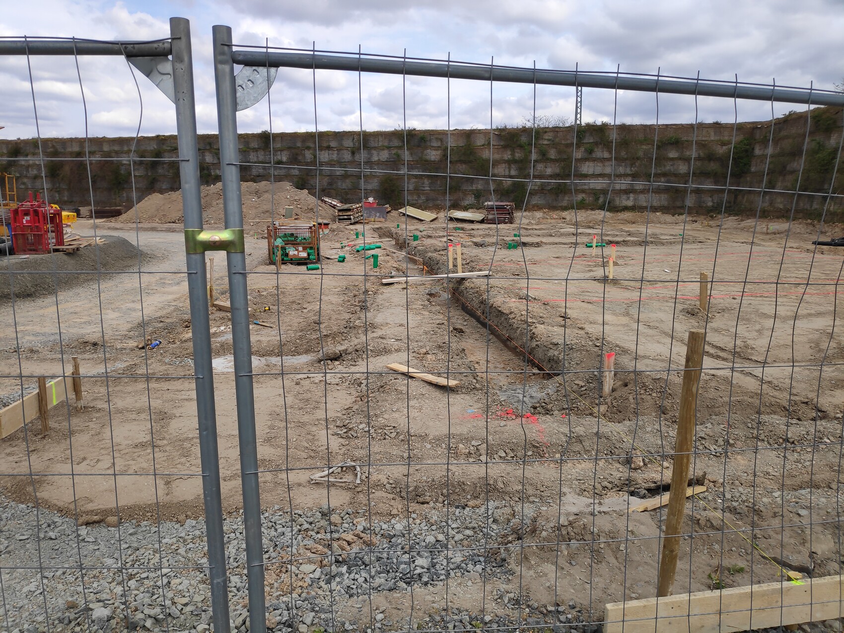 Baustelle hinter Drahtzaun: Graben, Baumaterialien, Holzpfähle, Zugangsbereich, Wolkenhimmel.