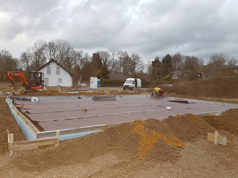Fundamentplatte auf Baustelle; Bagger rechts, zwei Arbeiter am Aufbau; Häuser im Hintergrund
