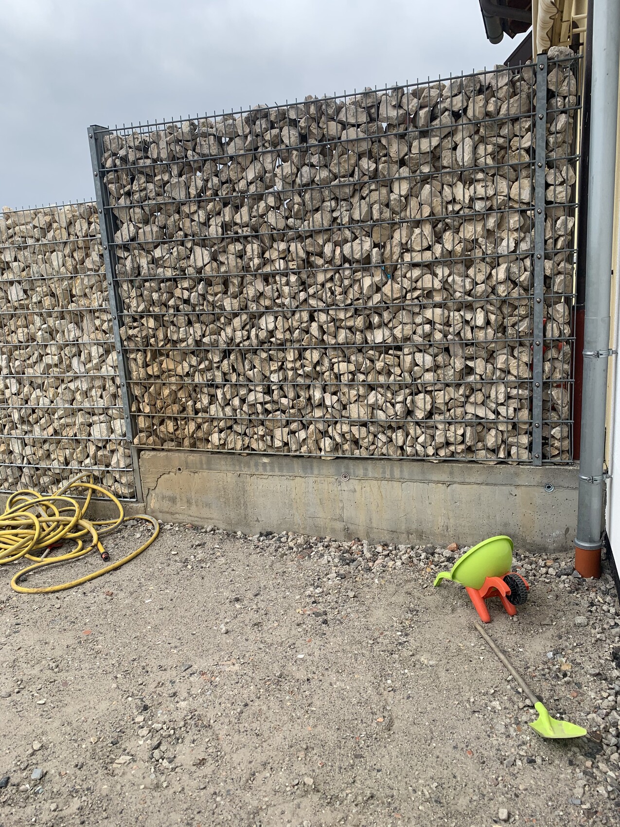 Gabionenwand aus Steinen vor Betonfundament; gelber Schlauch, buntes Spielzeug auf Kies.