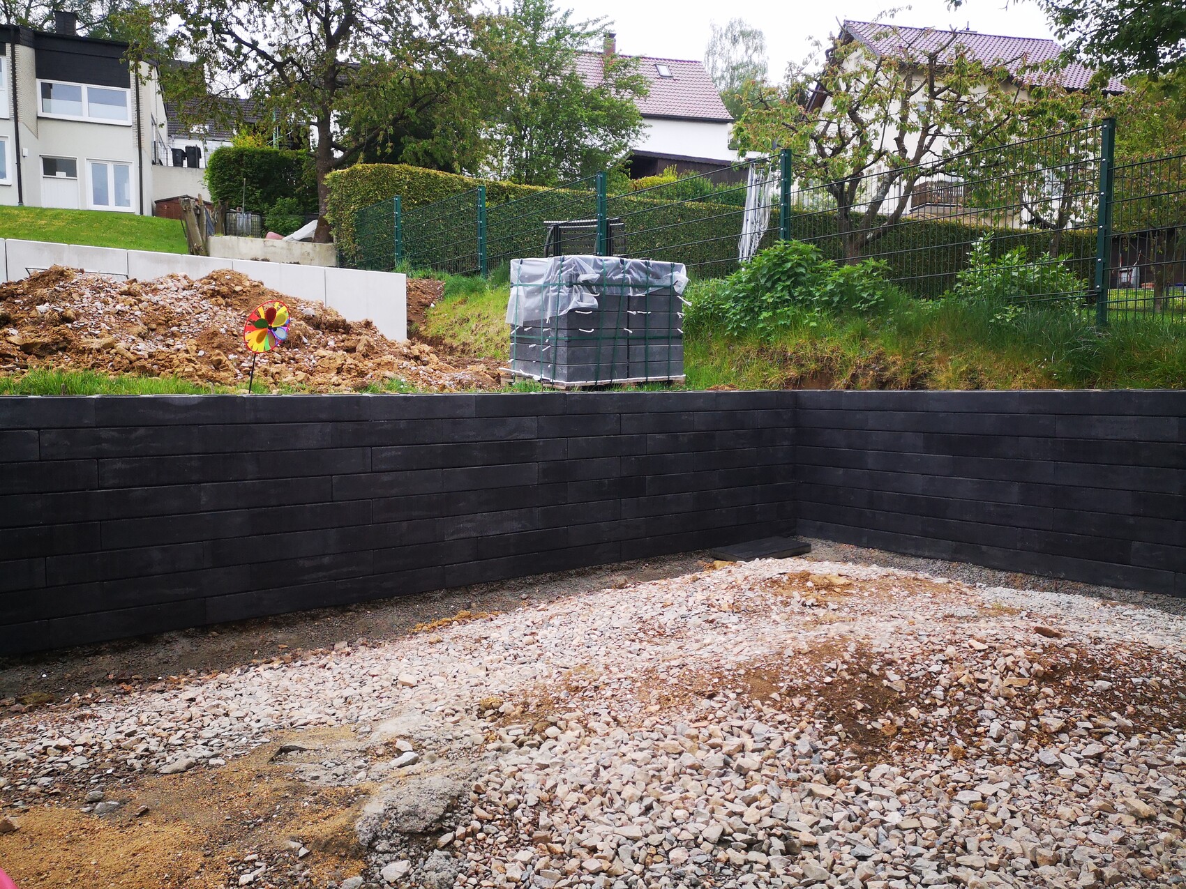 Baustellenszene im Garten: schwarze Holzplatten-Wand, Kiesboden, Erdhügel, bunte Windmühle.