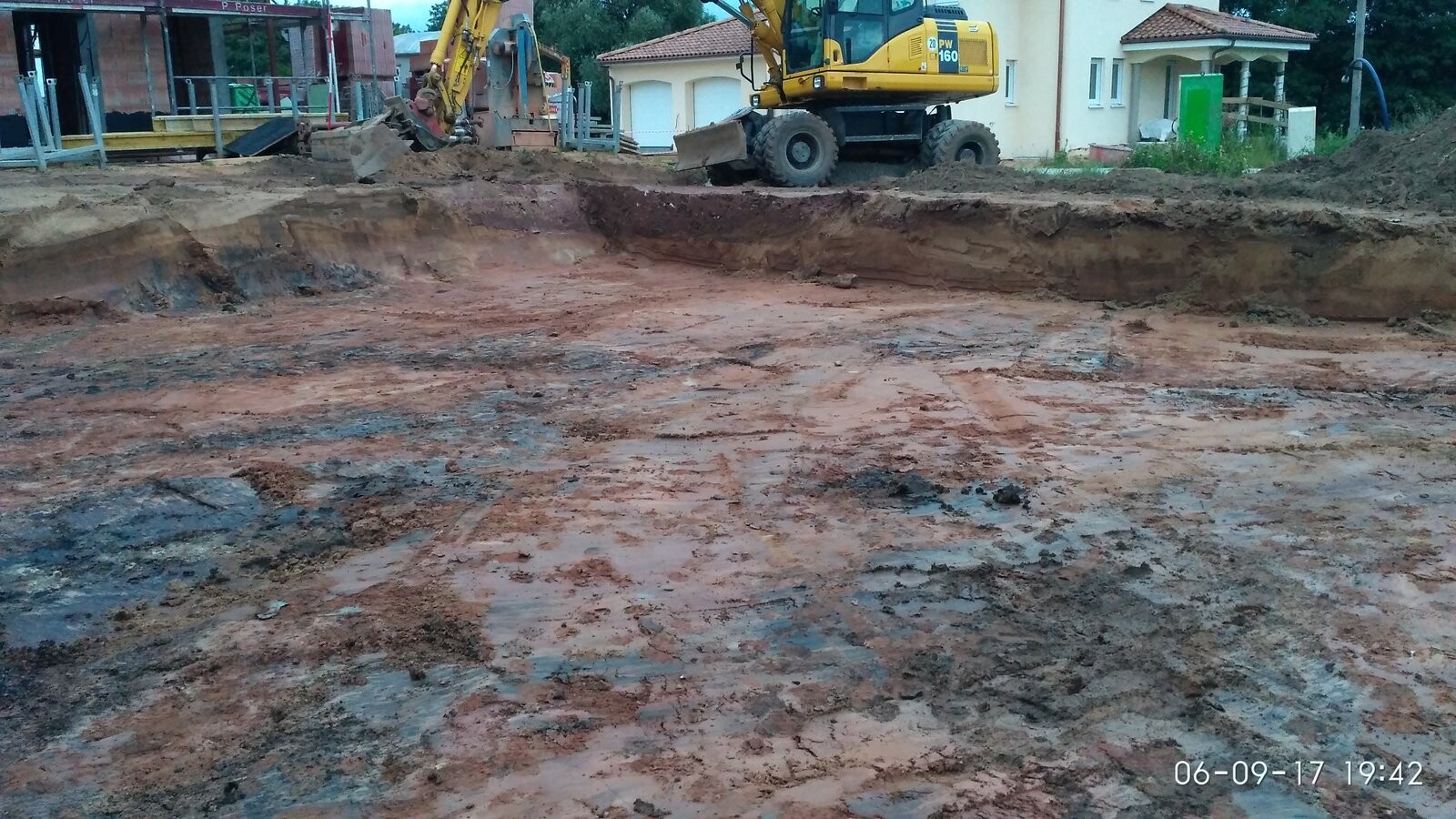 Baustelle mit gelbem Bagger, tiefe Baugrube und feuchte Erde, Häuser im Hintergrund.