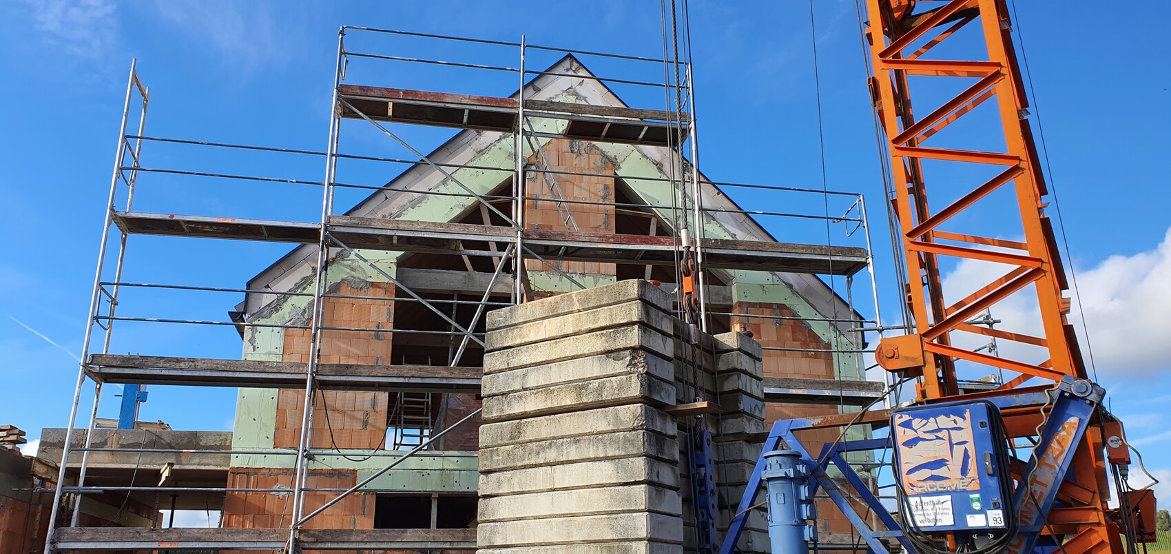 Baustelle mit Gerüst um halbfertiges Haus; Betonplatten vorne, oranger Kran rechts, blauer Himmel.