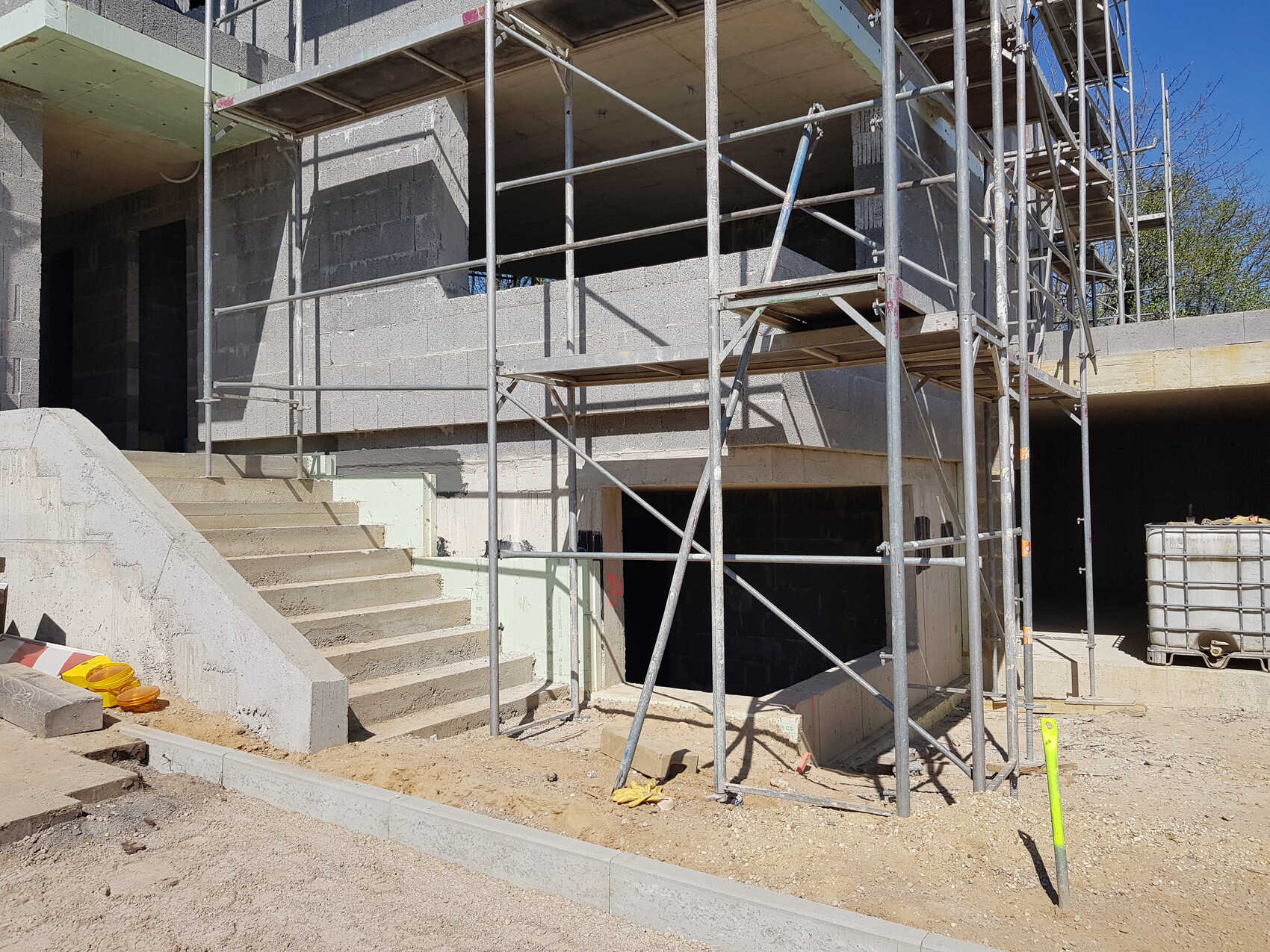 Baustelle mit Gerüst um ein unfertiges Betongebäude, Treppenstufen und Baucontainer