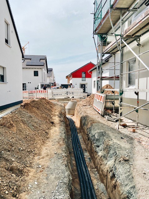 Baustelle: Baugrube mit schwarzen Rohren, Absperrungen, Gerüst an Gebäuden.