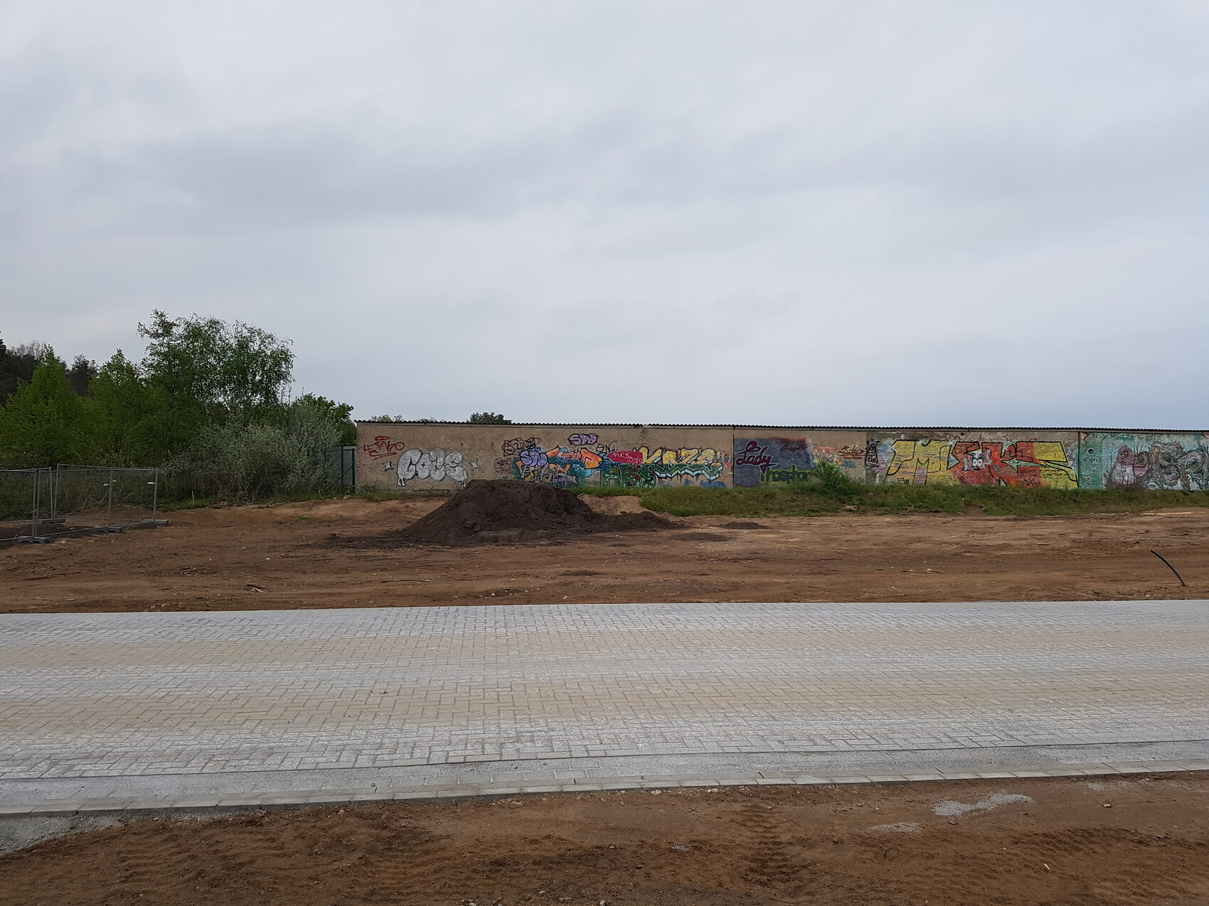 Offene Baustelle: Erdhügel, graffiti-bemalte Längswand, gepflasterter Vorplatz, wolkiger Himmel.