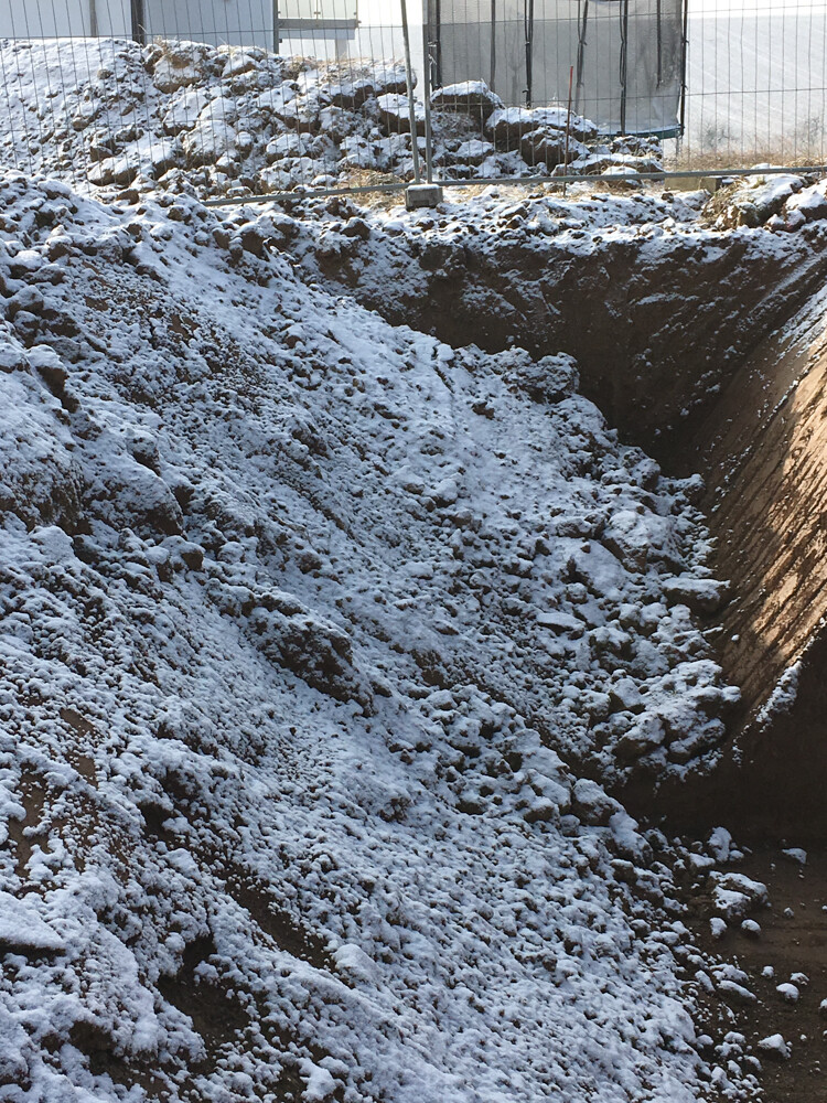 Baustellen-Grube mit Schnee und Erdaushub hinter Zaun
