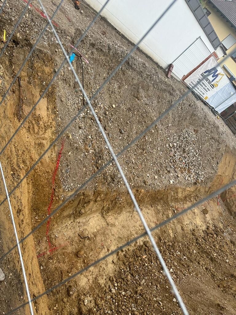 Baugrube auf einer Baustelle hinter Bauzaun, Erd- und Kiesboden, Gebäude im Hintergrund